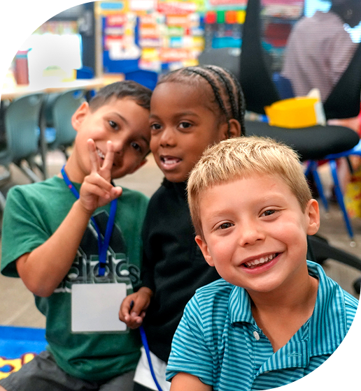 Three children smiling and posing together in a colorful classroom setting.