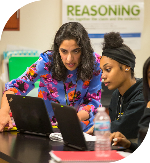 A woman in a floral dress assists students working on laptops in a classroom setting.