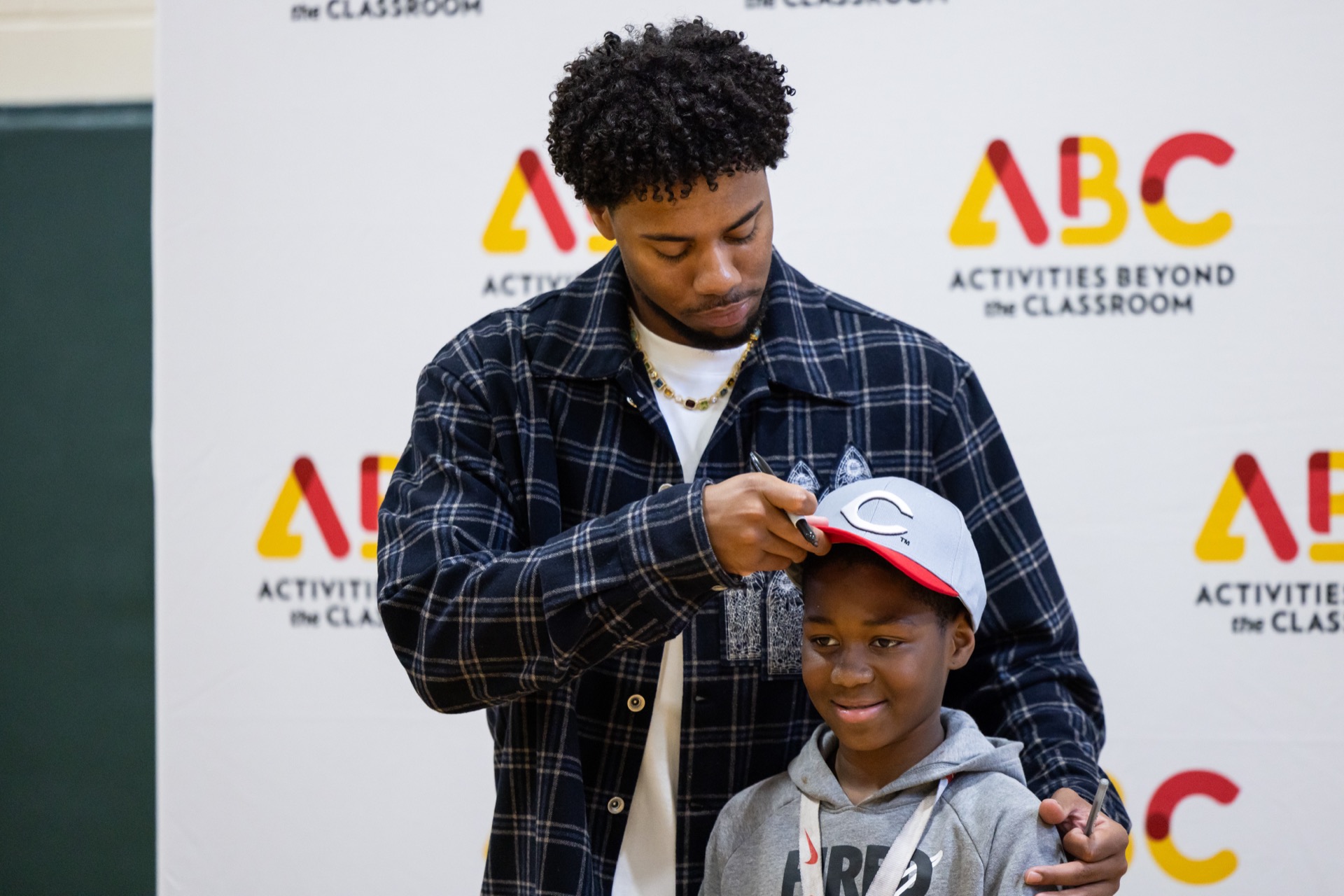 A person is adjusting a cap on someone else's head in front of a backdrop with a logo.