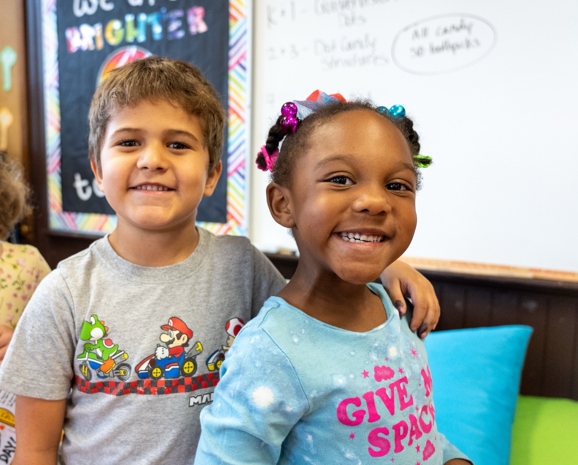 Two children smiling in a classroom setting with colorful decor and a welcoming atmosphere.