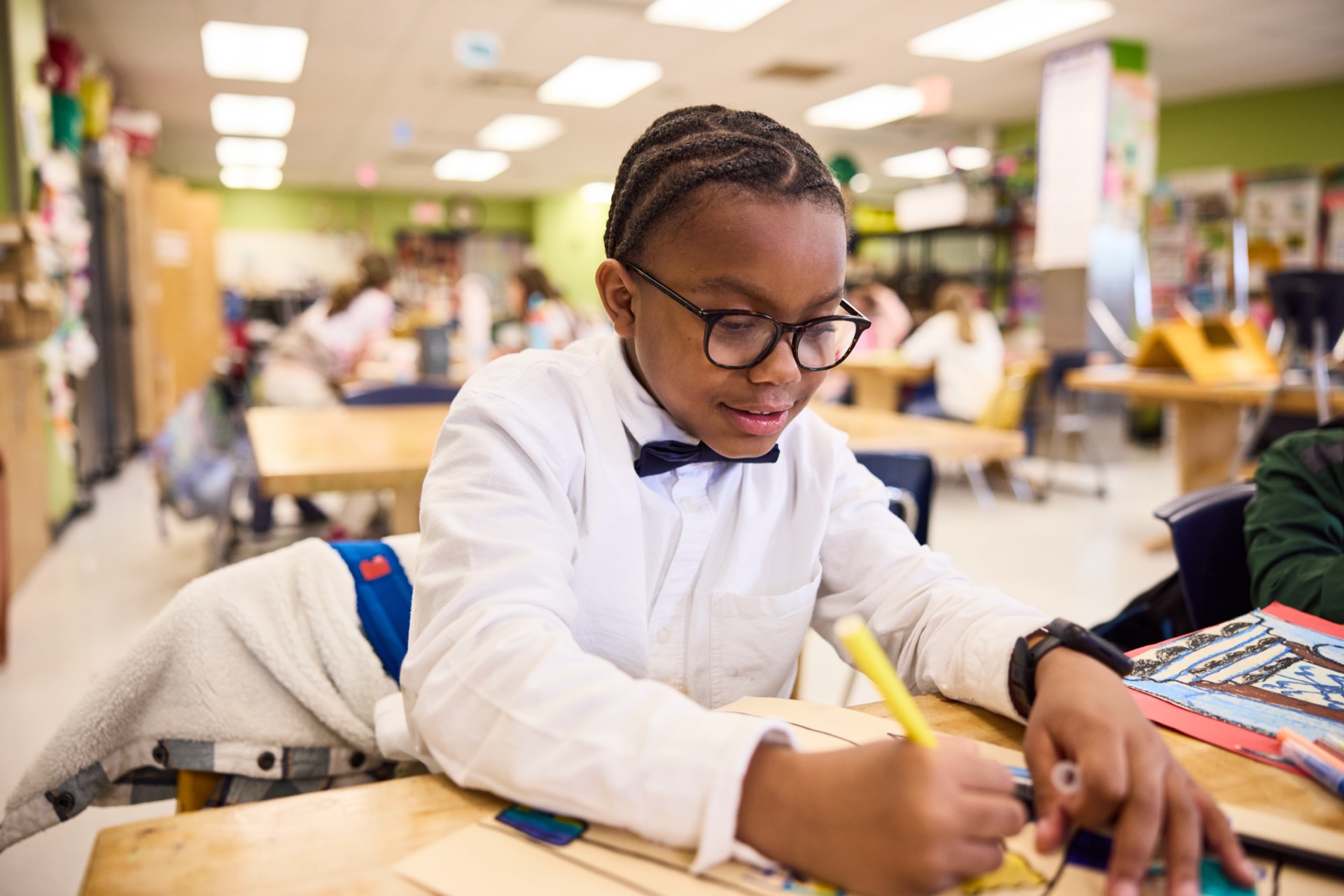 A student with glasses, wearing a white shirt and bow tie, is focused on writing at a classroom desk.