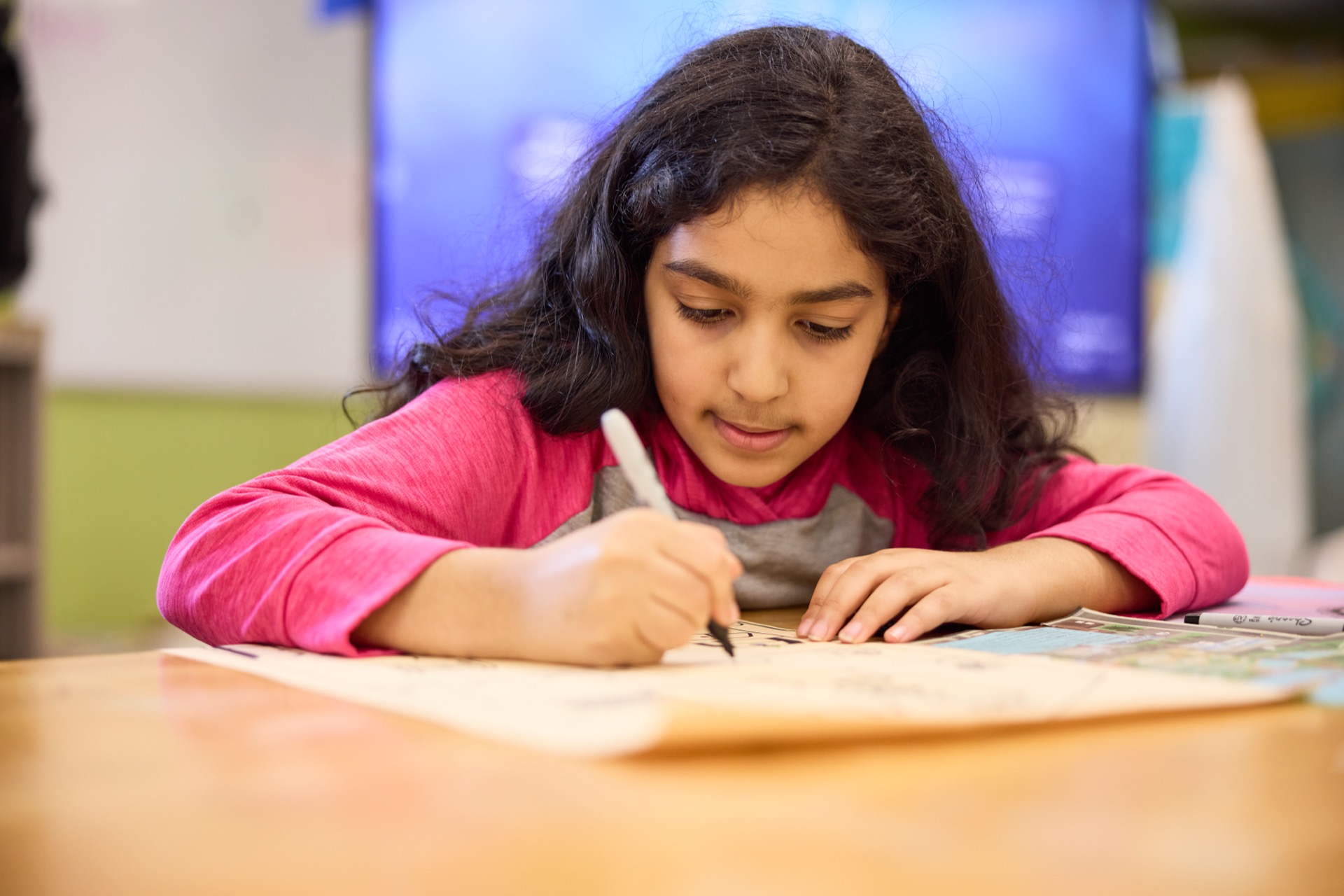 A young girl with long hair is drawing on a sheet of paper while wearing a pink long-sleeve shirt.