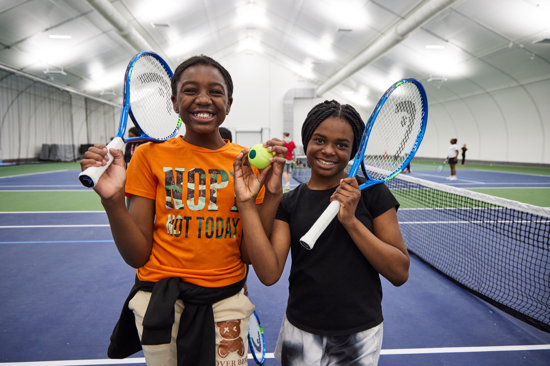 Two people smiling and holding tennis rackets and balls in an indoor tennis court.