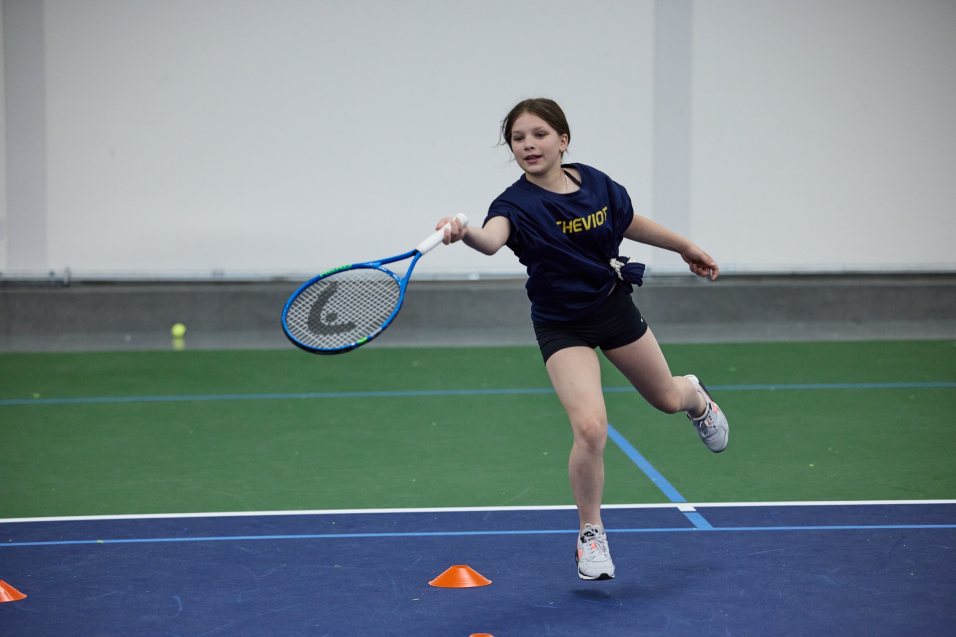 A person in athletic clothing is playing tennis indoors, reaching for a forehand shot near orange cones.