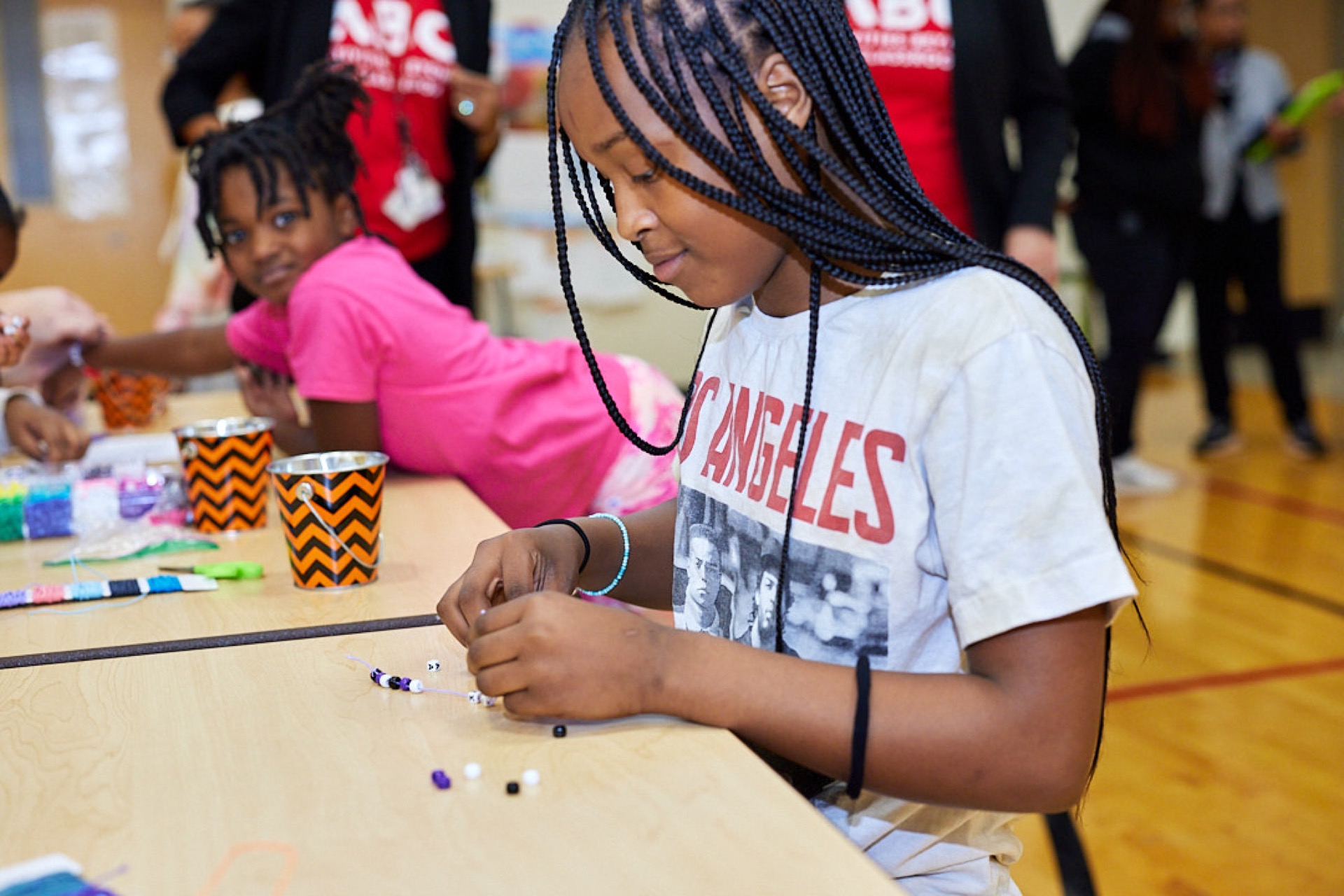 A young girl is focused on threading beads on a string at a wooden table, with another child partially visible in the background.