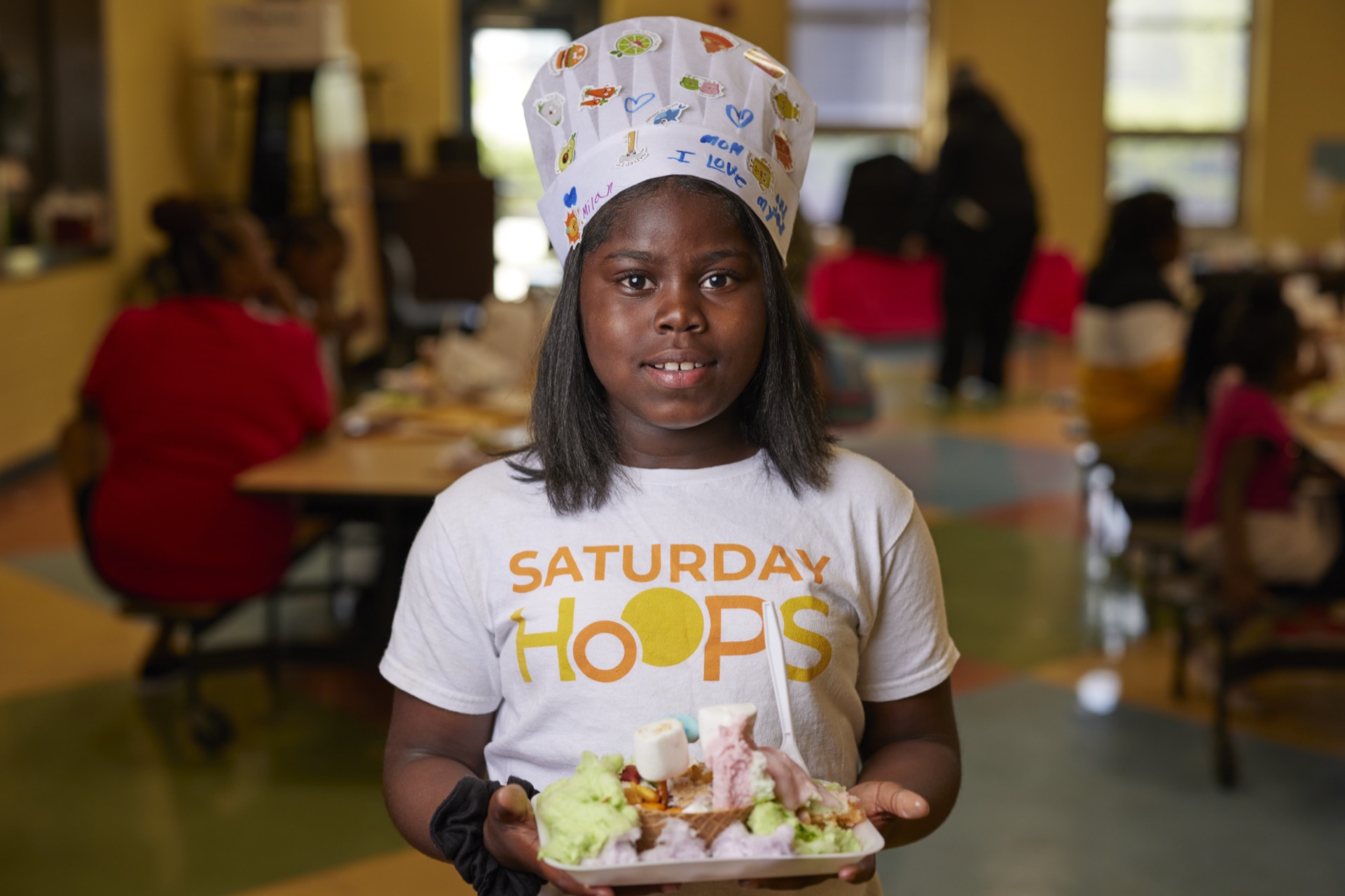 A person wearing a "Saturday Hoops" shirt and a decorated chef's hat holds a plate of food.