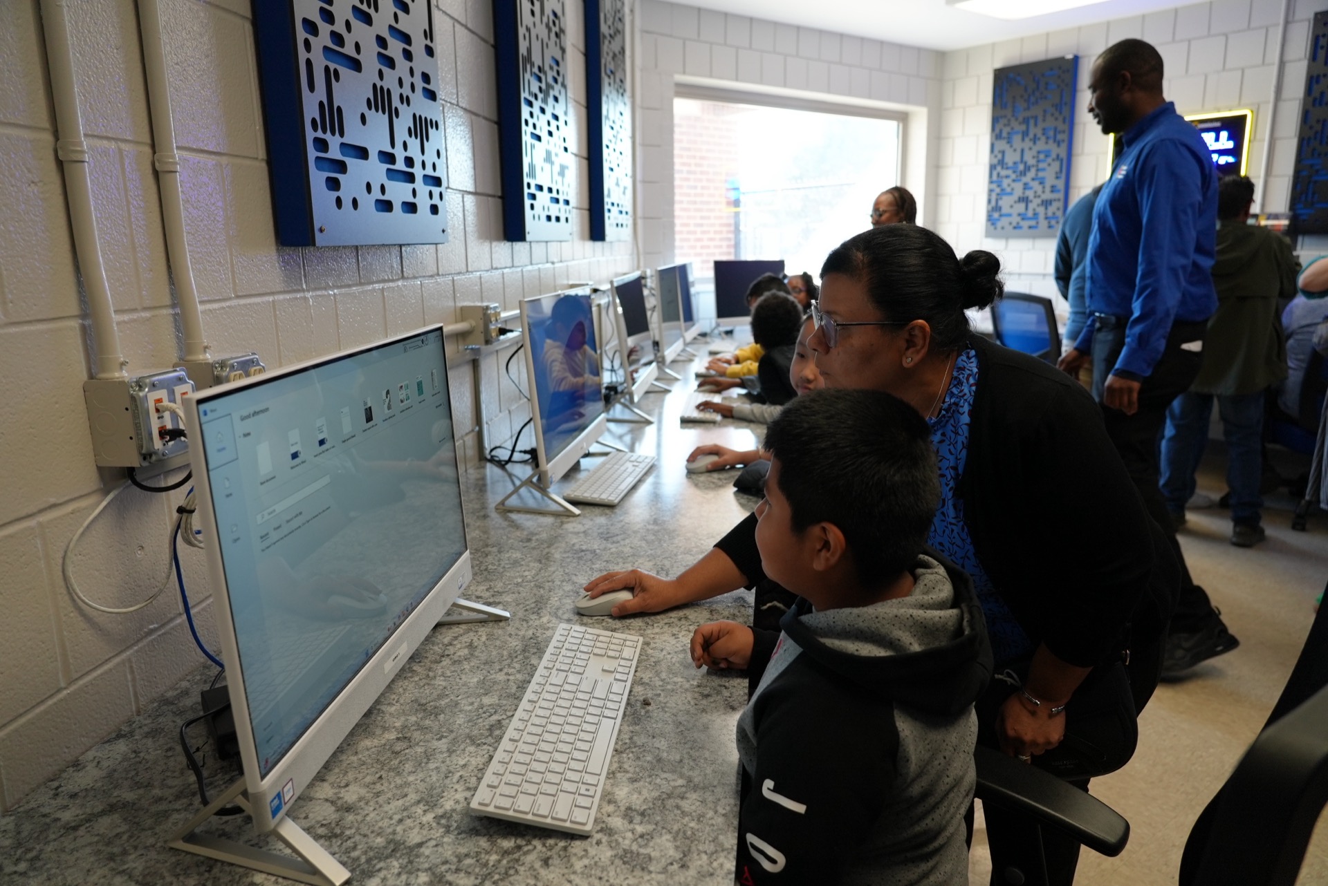 In a computer lab, people are actively working on desktop computers with guidance from an instructor.