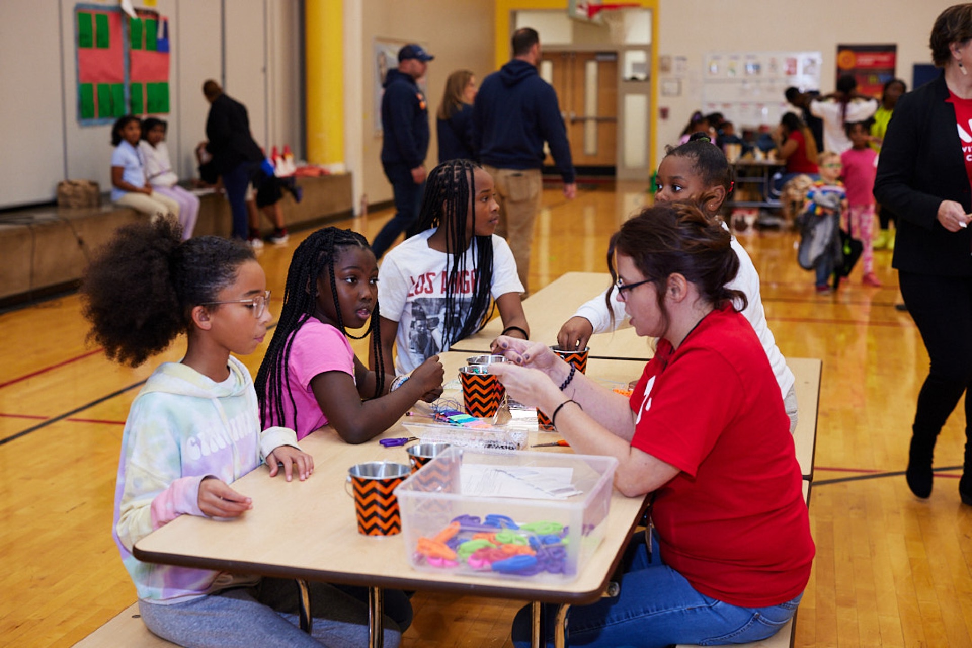 A group of children sits around a table engaging in crafts with an adult in a school gymnasium.