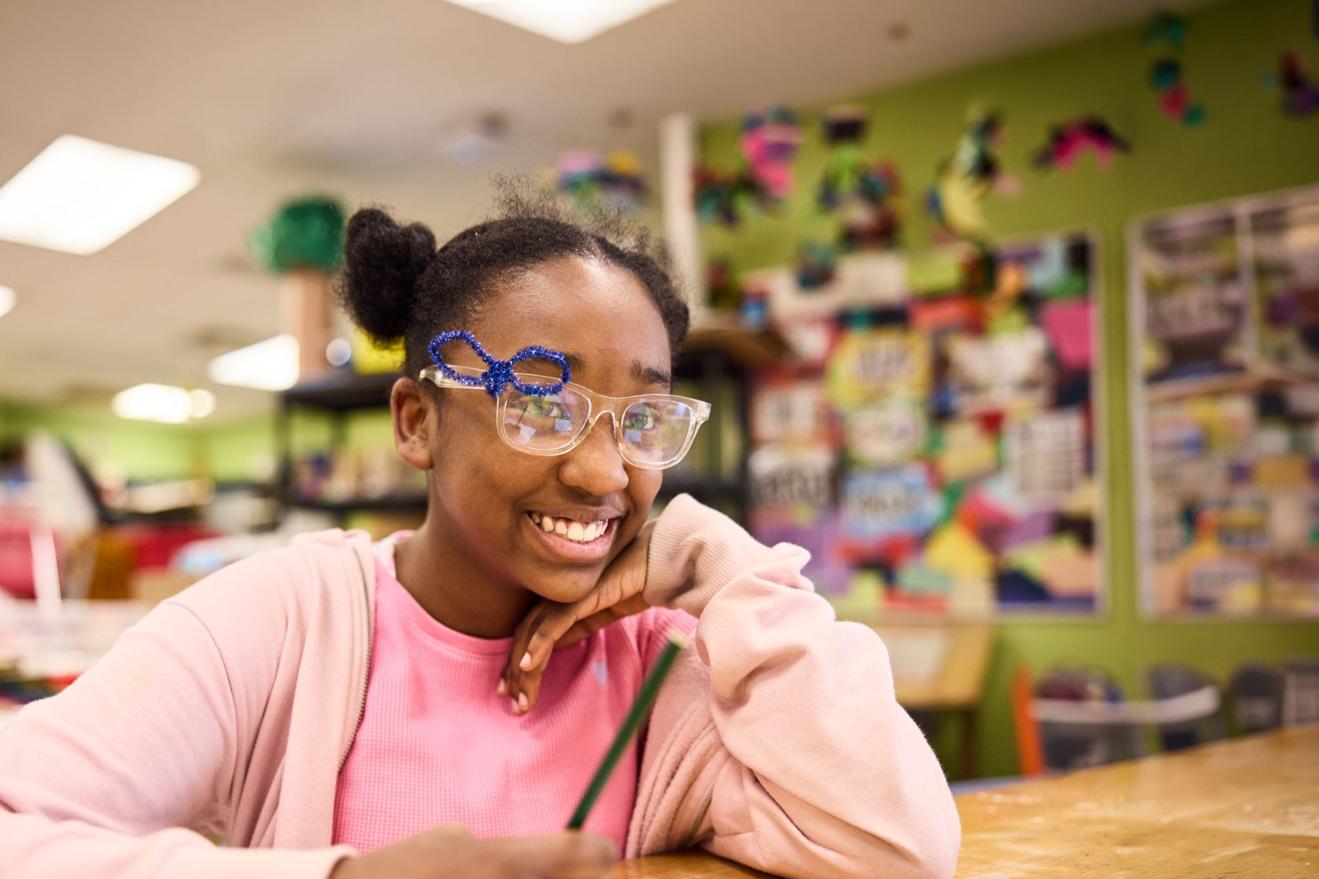 A person wearing safety goggles and a pink sweater smiles while sitting at a desk, holding a pencil.